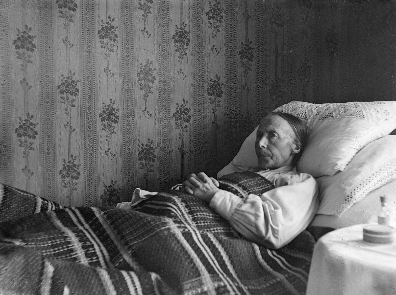 An elderly person lies in bed, covered by a blanket.